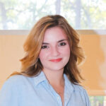 amber willburn Young blonde woman in a light blue shirt and white pants stands indoors by a wooden counter, large windows and trees behind her.