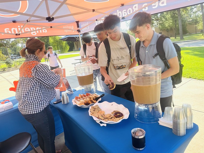 Students gather at an outdoor table with pastries as a woman serves drinks from dispensers under a canopy on a sunny day.
