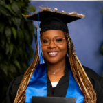 _Artavia Davis AC Grad AAT Testimonials Smiling graduate with a decorated cap, glasses, and blue stole holds a diploma before greenery and a blue wall.