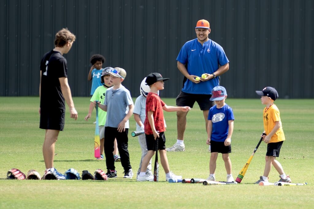 Children with baseball bats chat and laugh on a grassy field as two adults supervise; gloves rest nearby on the grass.