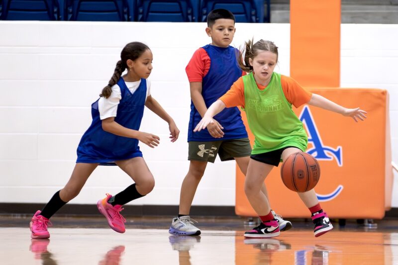 Three children play basketball indoors; a girl in an orange shirt dribbles while two in blue jerseys defend her.