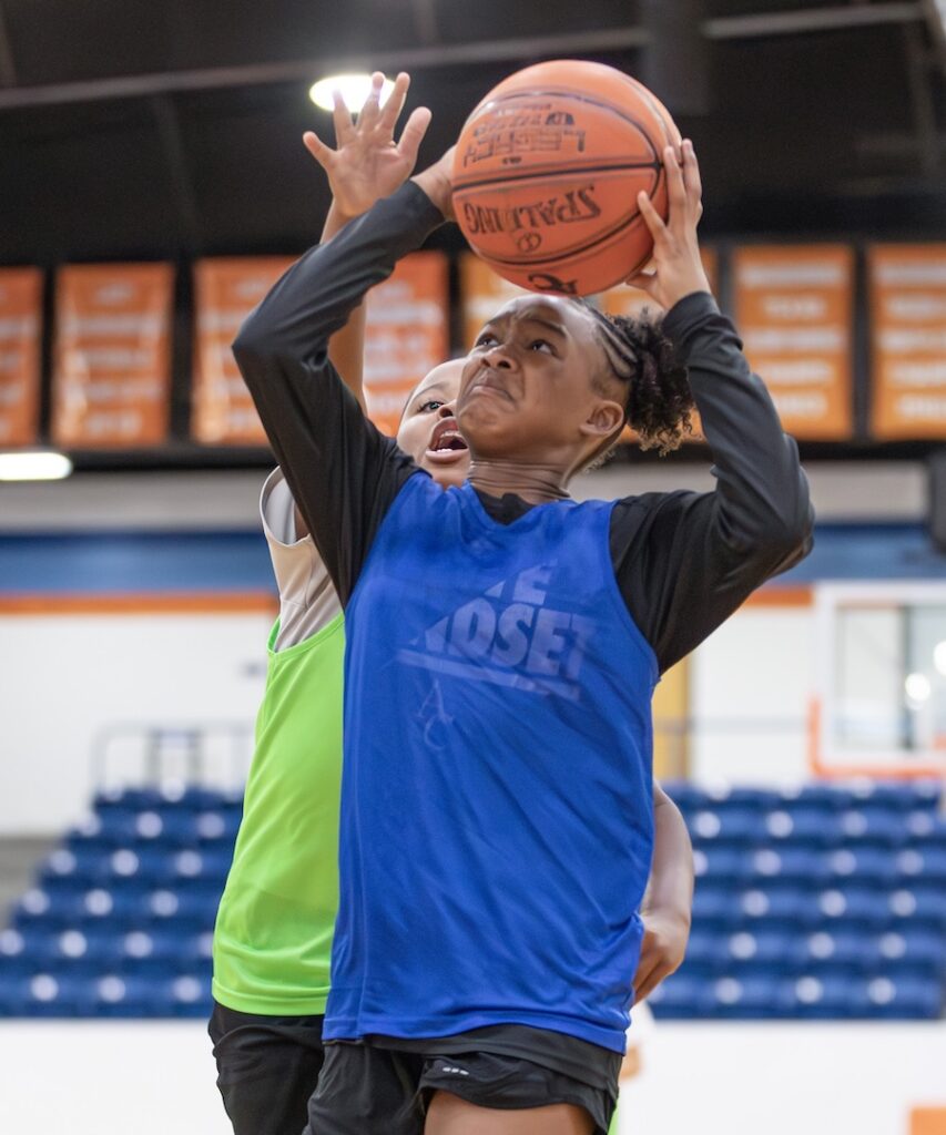 Player in blue jersey jumps to shoot as player in green jersey closely defends from behind during a basketball game.