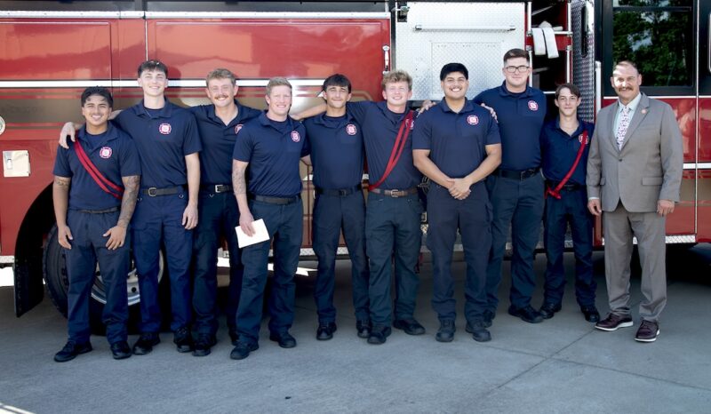 Eight people in navy uniforms smile before a red fire truck; a man in a tan suit stands at the far right.