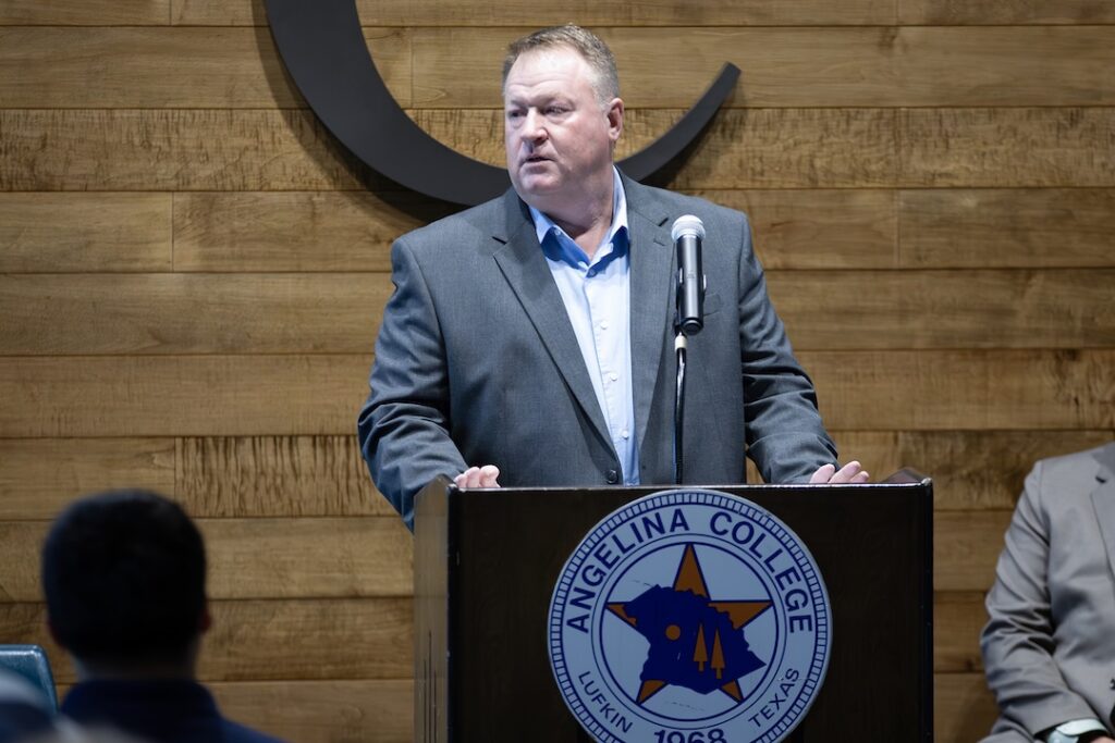 Man in gray suit speaks at podium with Angelina College logo; wooden wall background, another person partially visible right.