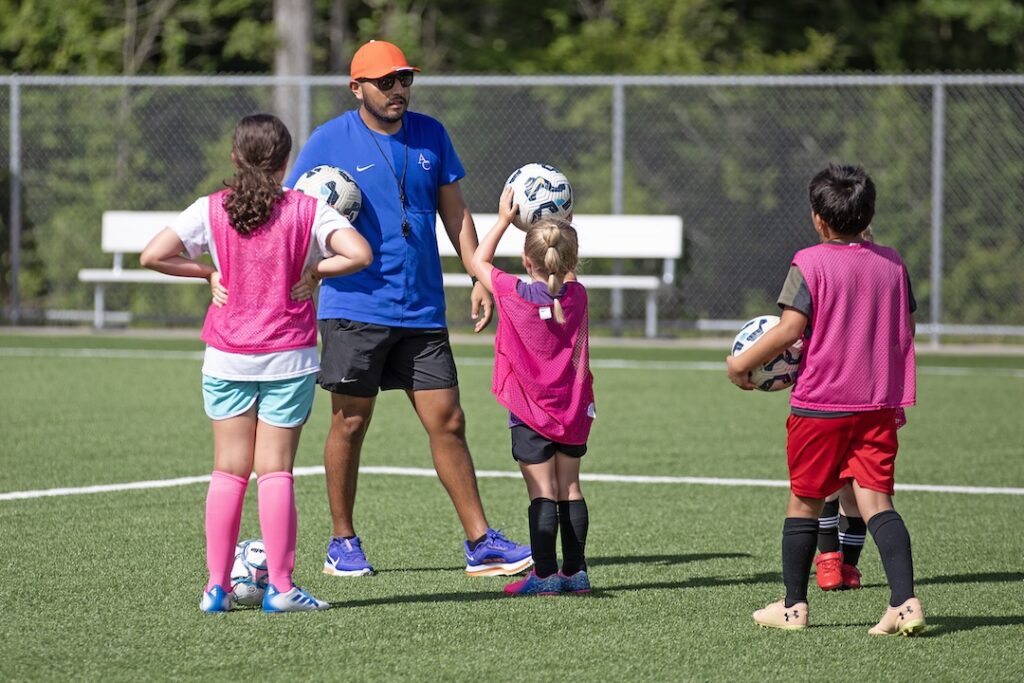 Soccer coach in sunglasses and orange cap talks to three kids in pink pinnies holding balls on an outdoor field.