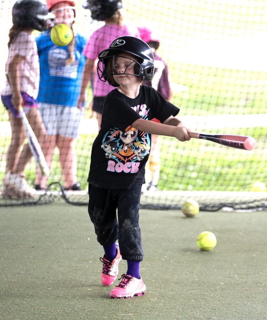 Child in helmet and black T-shirt swings at a baseball during practice, with other kids and balls near a netted area.