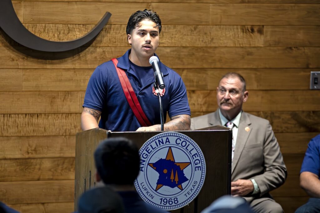 Young man speaks at Angelina College podium; older man in suit sits behind. Wooden wall and audience visible indoors.