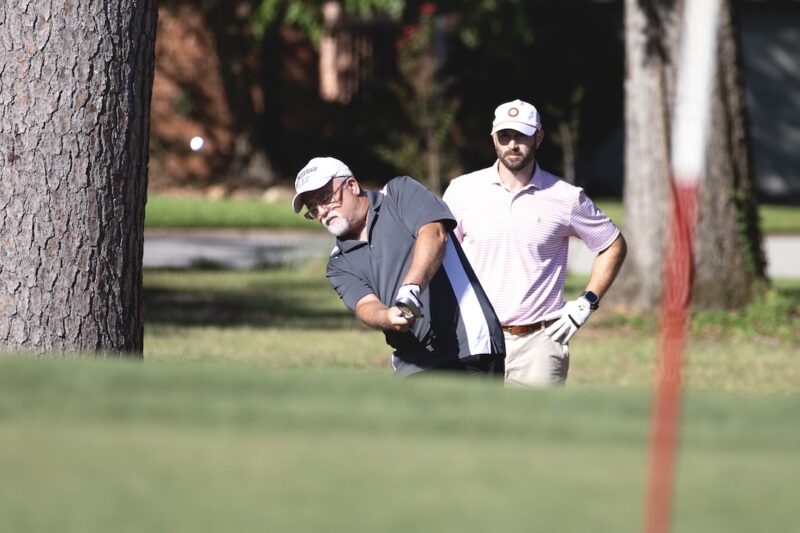 Two men play golf outside; one swings a club while the other watches. Trees and grass in the background on a sunny day.