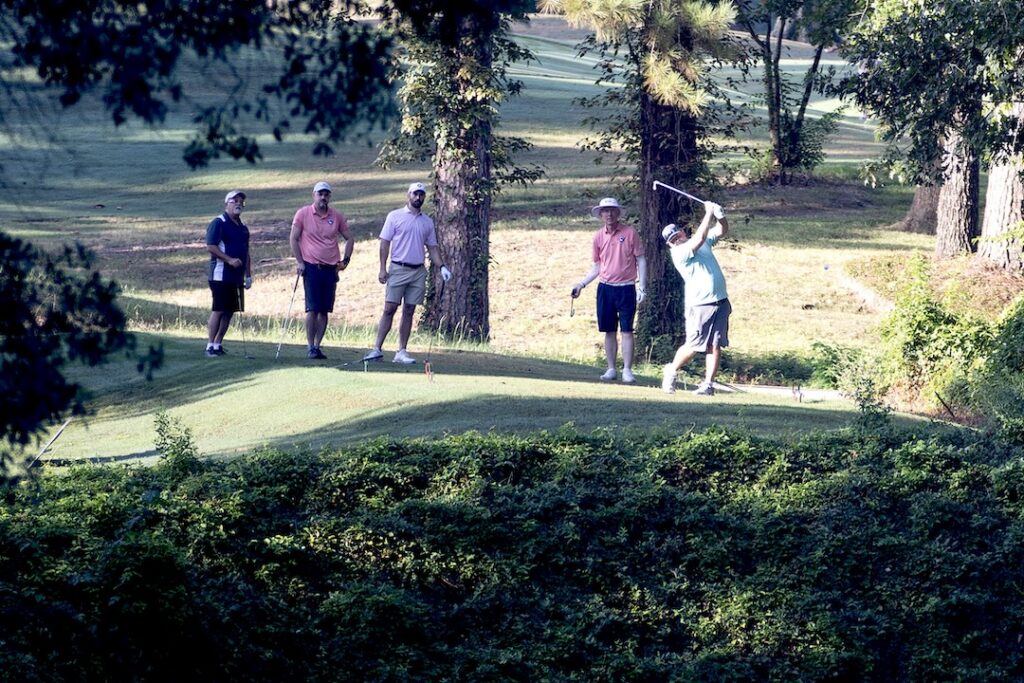 Four men on a sunny golf course; one swings a club while three watch, surrounded by trees and greenery.
