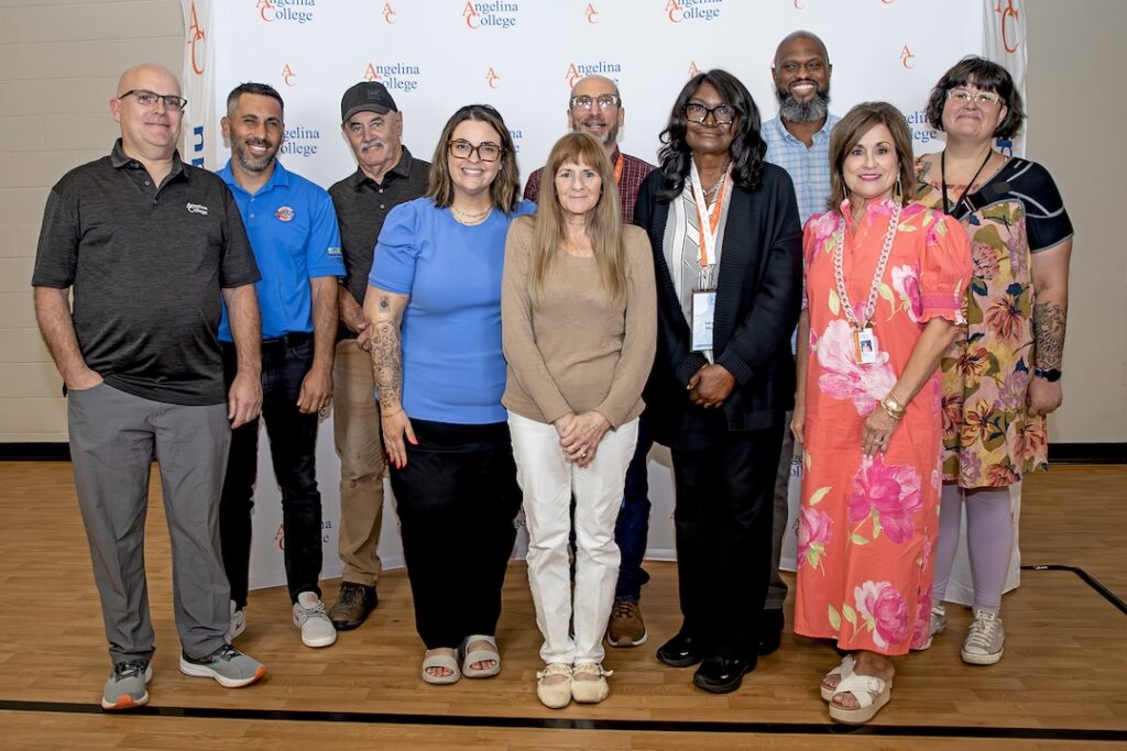 Eleven adults smile and pose indoors before an Angelina College backdrop, wearing business casual, colorful clothing.