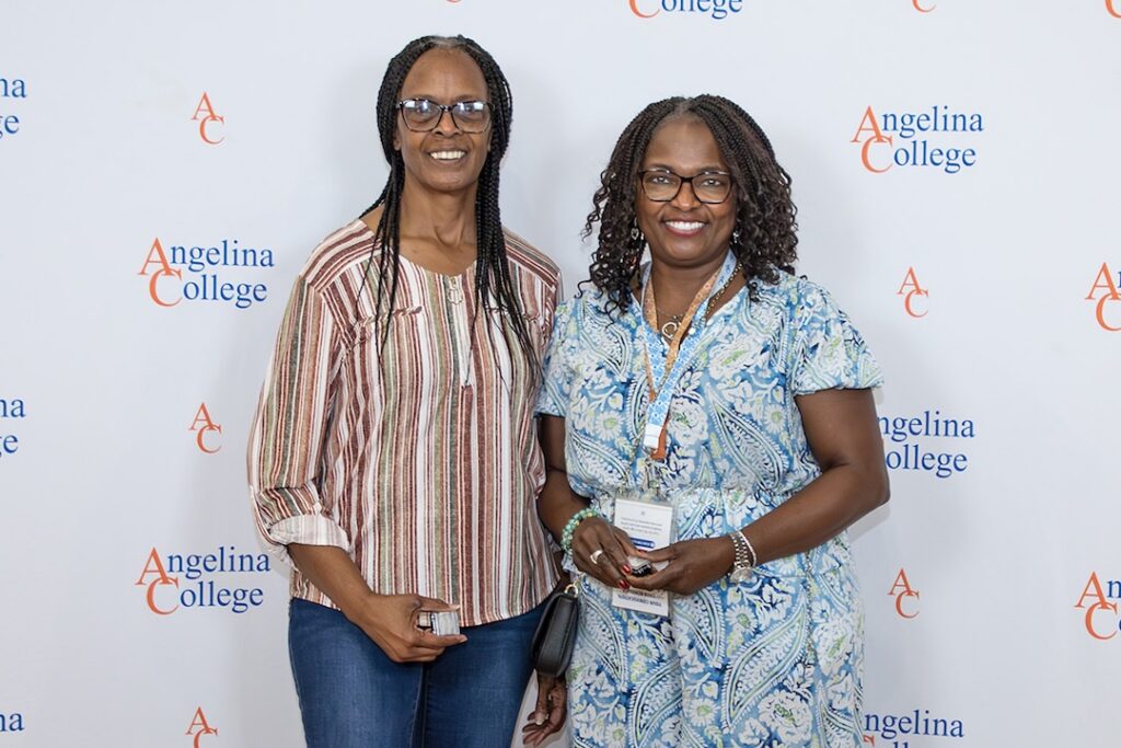 Two women wearing glasses and casual clothes smile in front of a step-and-repeat Angelina College logo banner.