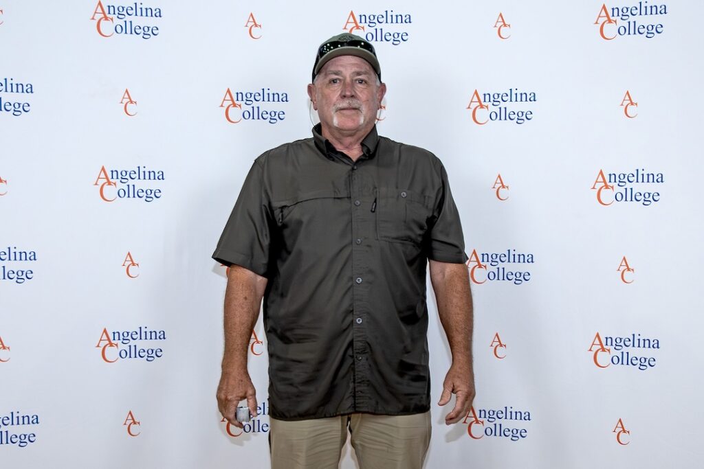 Man in dark short-sleeve shirt and cap stands before a banner displaying Angelina College logos and AC letters.