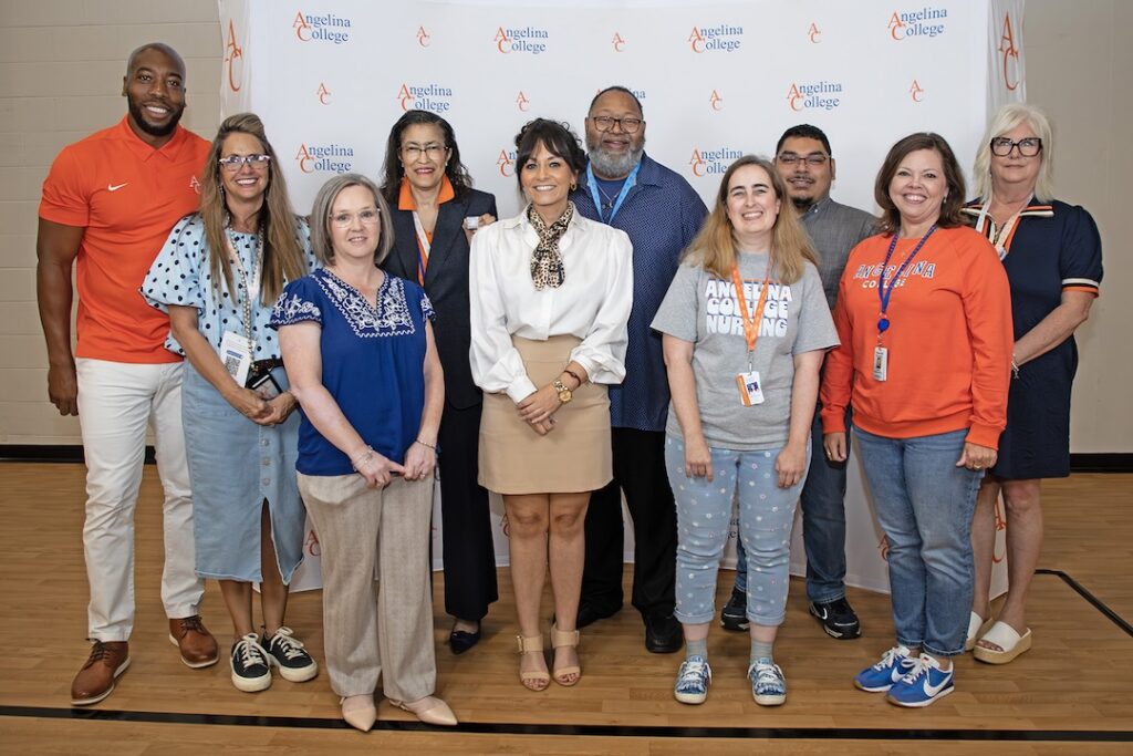 Eleven people smile on a gym floor in front of an Angelina College backdrop, dressed in casual and business attire.