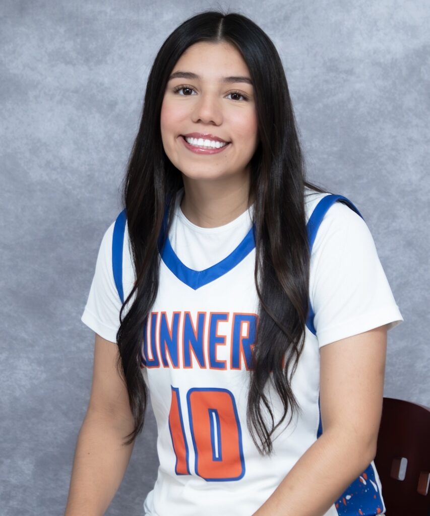 Young woman with long dark hair, smiling, wears a white and blue number 10 jersey while seated before a gray backdrop.