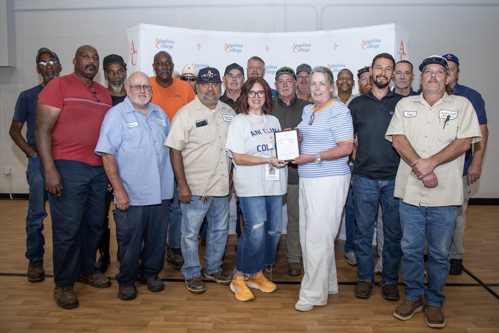 Seventeen adults, some in uniforms, pose on a gym floor as two women hold a certificate. Angelina College banner is behind them.