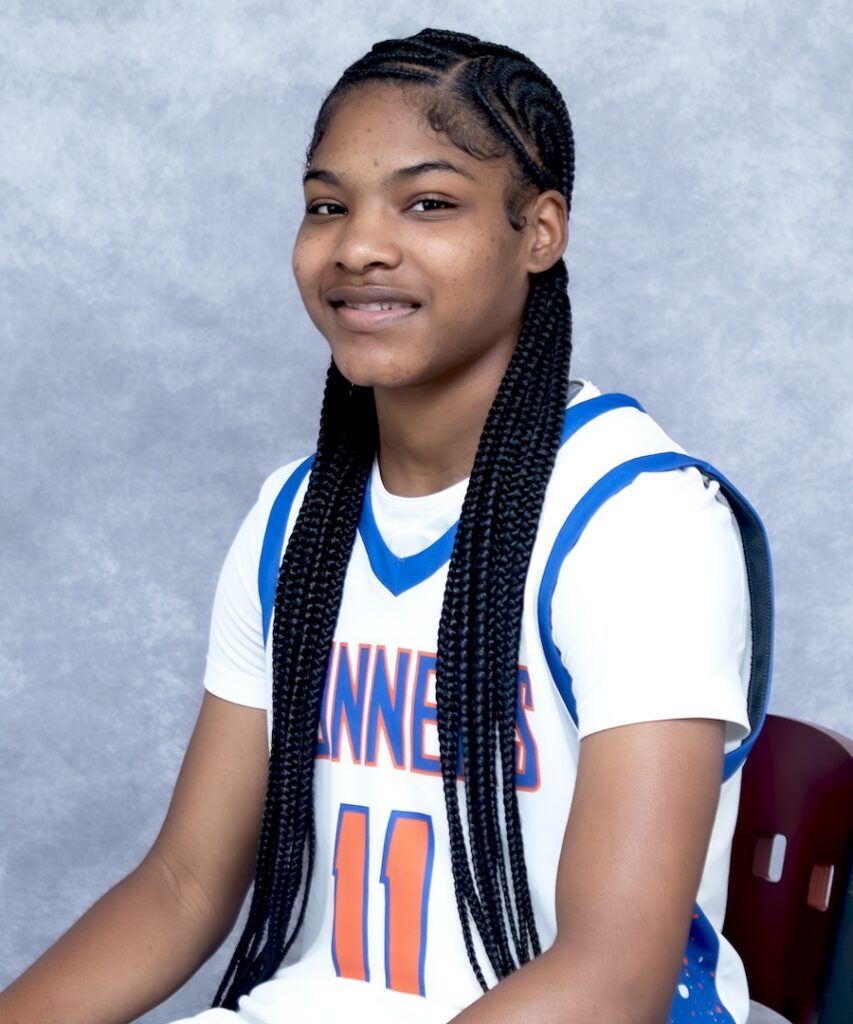 Young basketball player with long braids, in a white and blue number 11 jersey, smiles while seated before a gray backdrop.