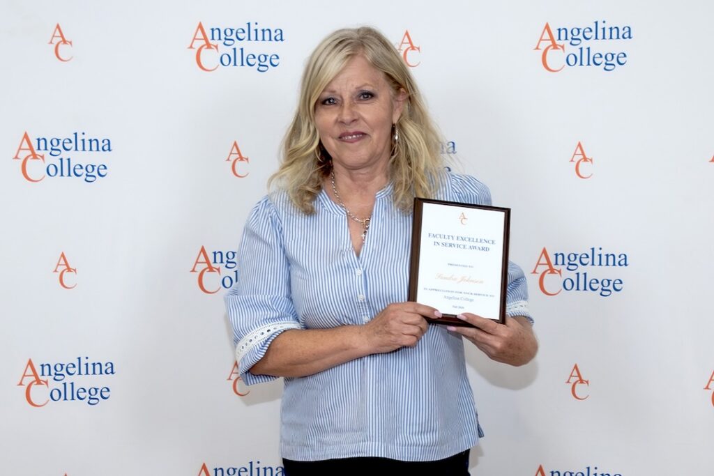 Smiling blonde woman holds a plaque in front of an Angelina College backdrop, wearing a blue and white striped blouse.