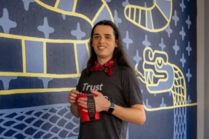 Person with long hair, red bandana, and smart watch holding a red cup in front of blue wall with yellow snake art.