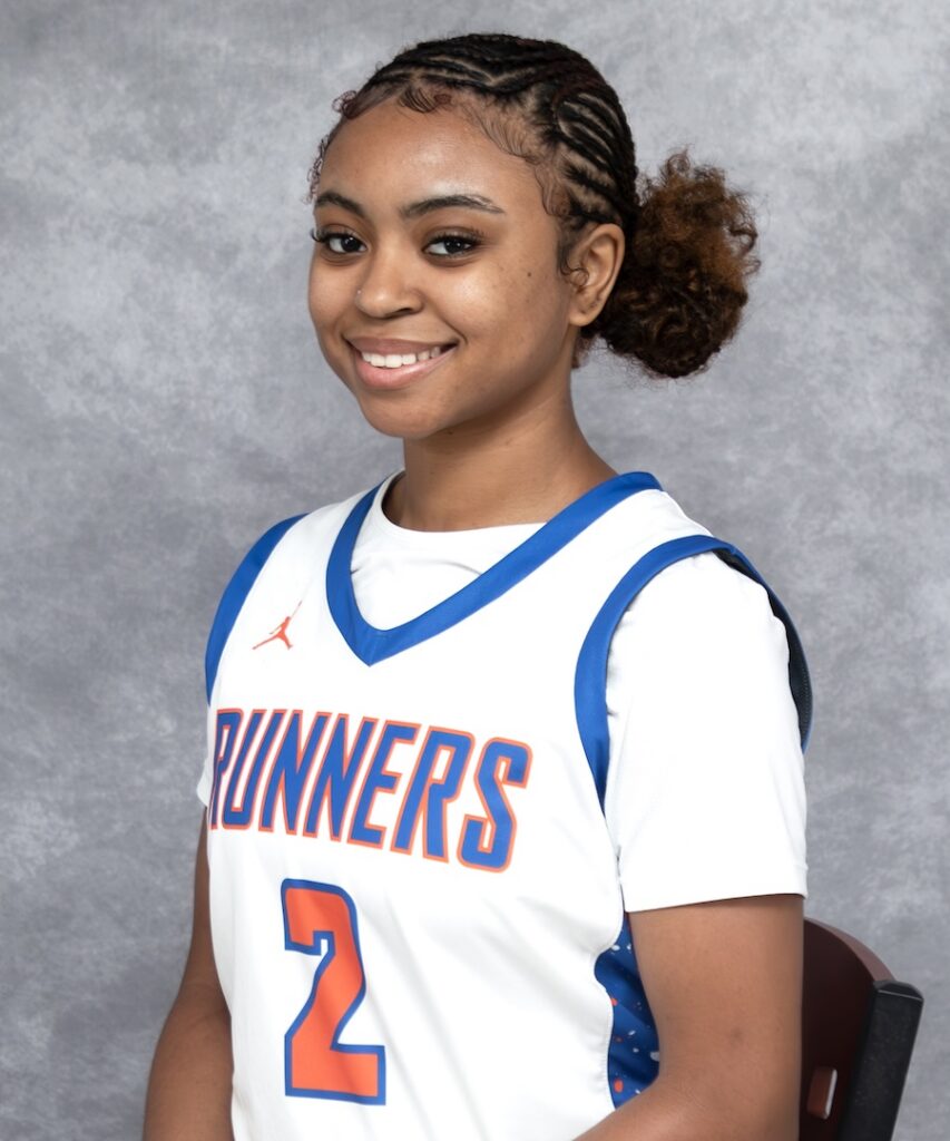 Smiling young woman in a white and blue Runners basketball jersey, number 2, seated against a gray background.