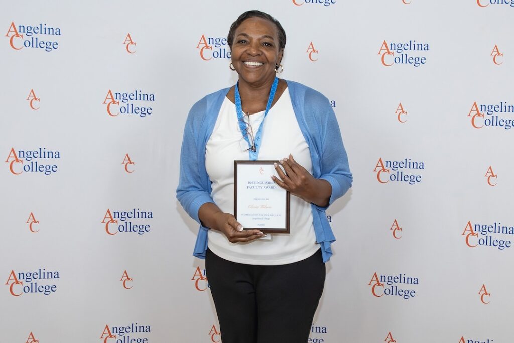 Woman in blue cardigan and lanyard smiles, holding a framed award before an Angelina College logo banner.
