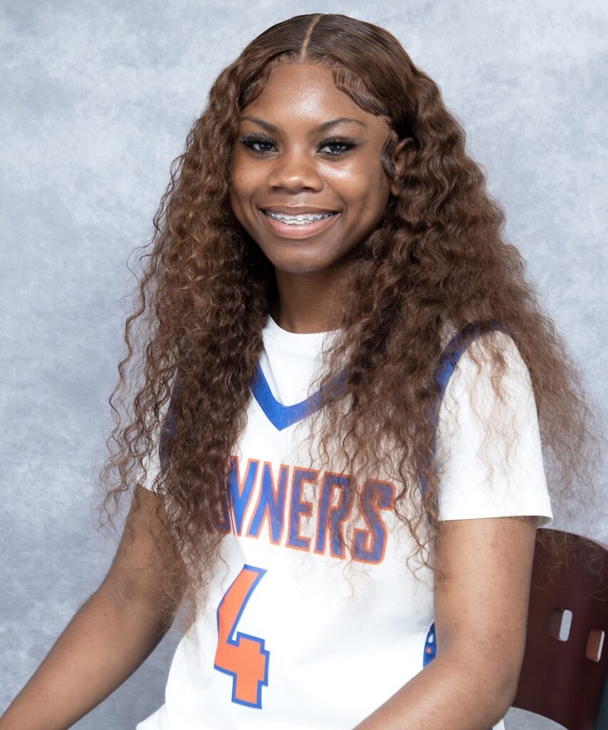 Young woman with long curly hair smiles while seated, wearing a white basketball jersey, number 4, against a light gray background.