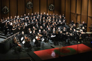 A choir in black stands behind seated orchestra musicians; wreaths decorate the back wall, instruments and stands at the front.