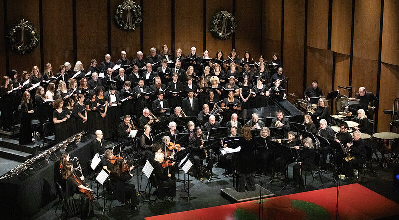 A choir in black stands behind seated orchestra musicians; wreaths decorate the back wall, instruments and stands at the front.