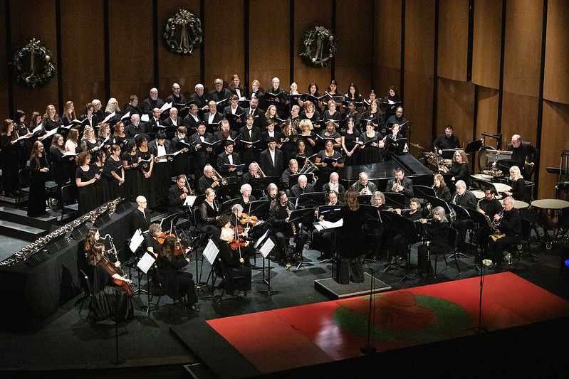 A choir in black stands behind seated orchestra musicians; wreaths decorate the back wall, instruments and stands at the front.