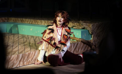 A person in dramatic stage makeup and a colorful shawl sits on a lit wooden floor, looking upward in thought.