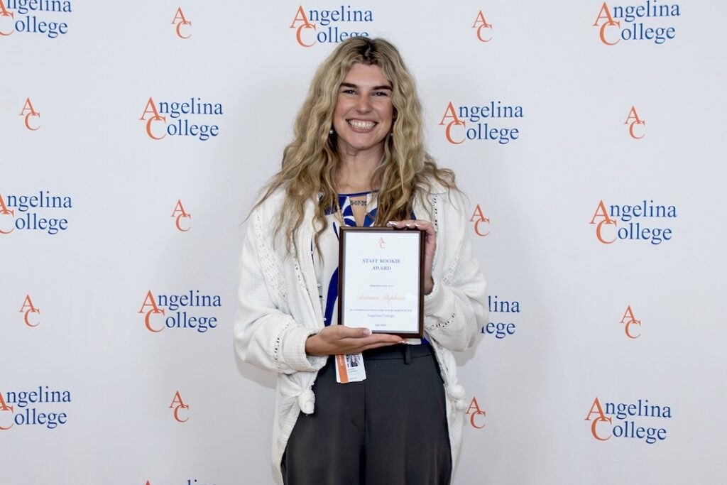Smiling person with long wavy hair holds a framed award in front of an Angelina College backdrop.