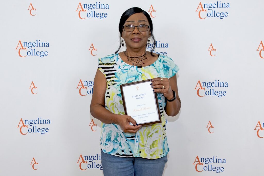 Woman in glasses and floral top smiles, holding a Staff Spirit Award before an Angelina College logo banner.