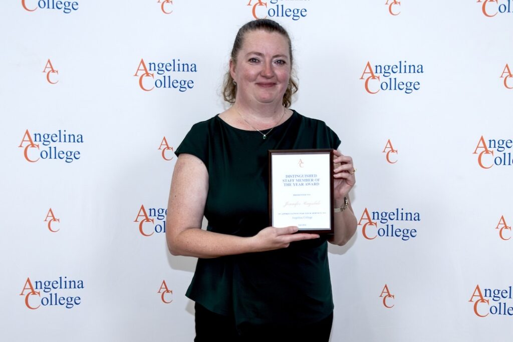 Smiling woman in a dark green top holds a Distinguished Staff Award plaque before an Angelina College backdrop.