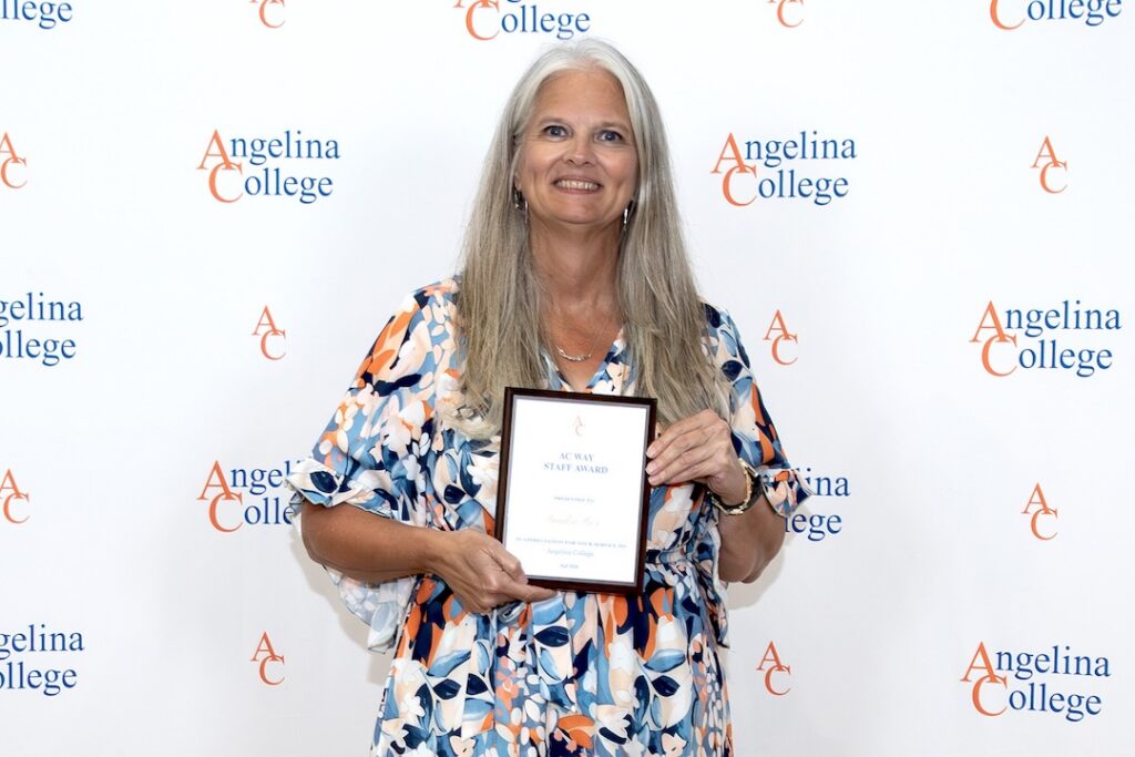 Woman with long gray hair in a floral dress smiles, holding a plaque in front of an Angelina College logo backdrop.