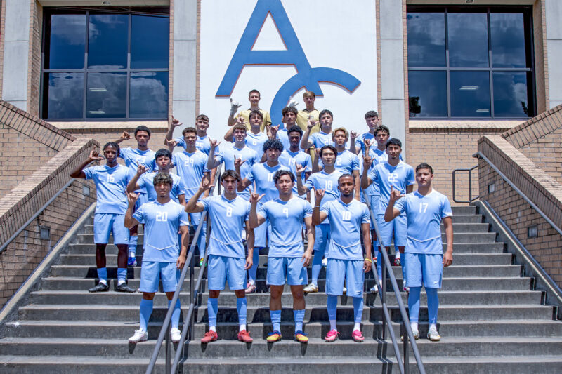 Soccer team in light blue uniforms poses on stairs, flashing peace signs in front of a building with large windows and A C logo.