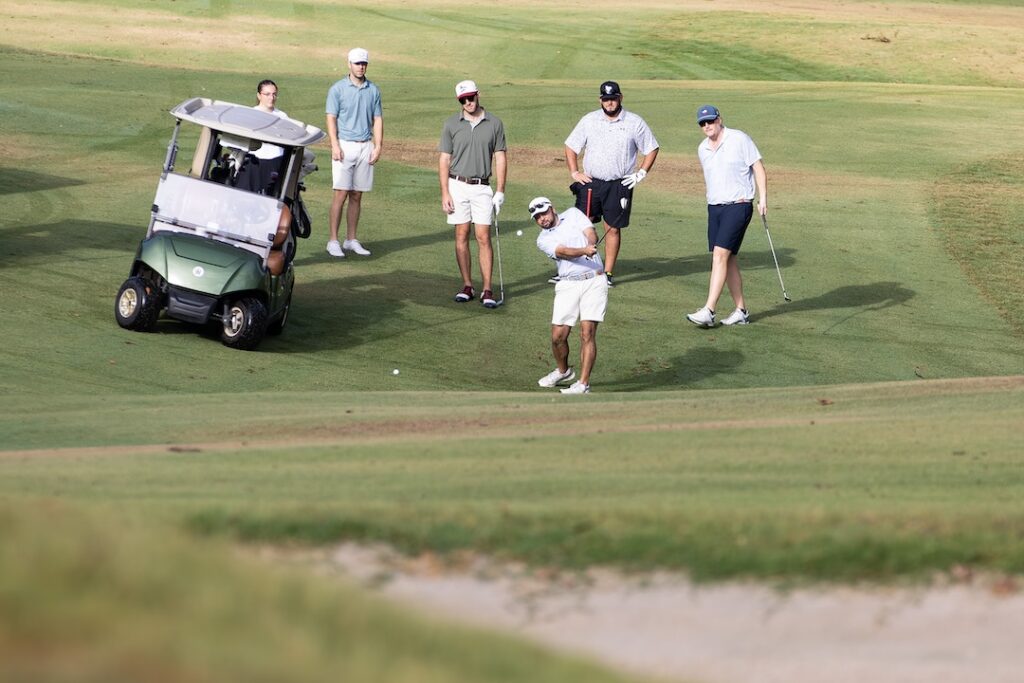 Six men on a sunny golf course; one swings a club as five watch. A green golf cart is parked on the grass nearby.