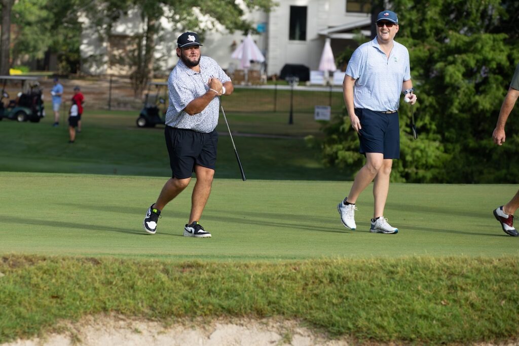 Two men in shorts and caps on a golf course; one swings a club, the other watches and smiles. Trees, building, and carts behind.