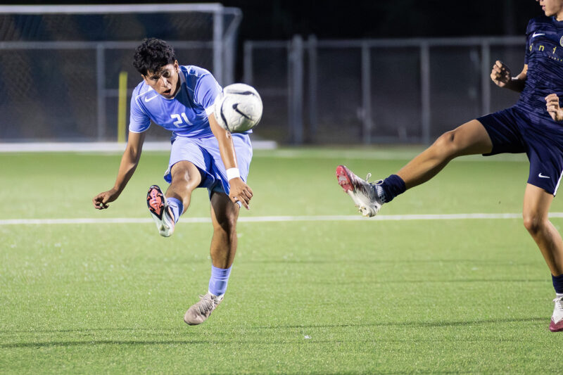 Soccer player in blue uniform jumps to kick the ball midair as another player’s leg reaches toward it on a green field.