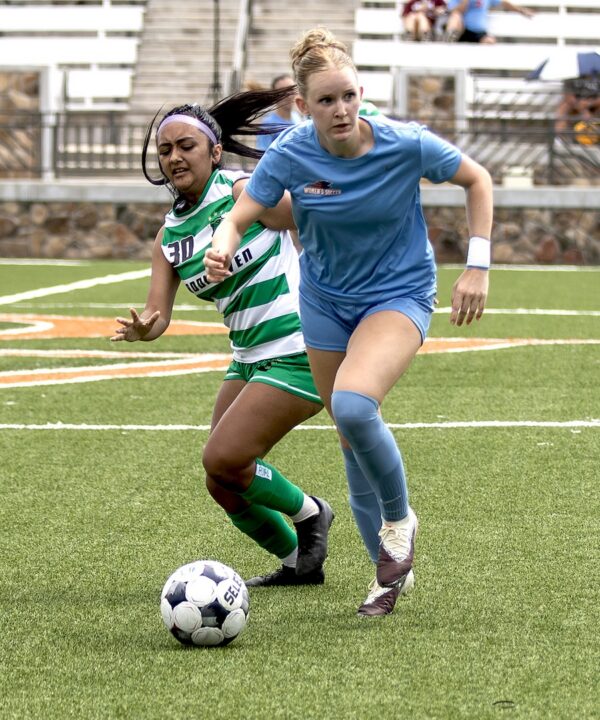 Two female soccer players, one in green and white, one in blue, compete for the ball on a field with empty bleachers behind them.