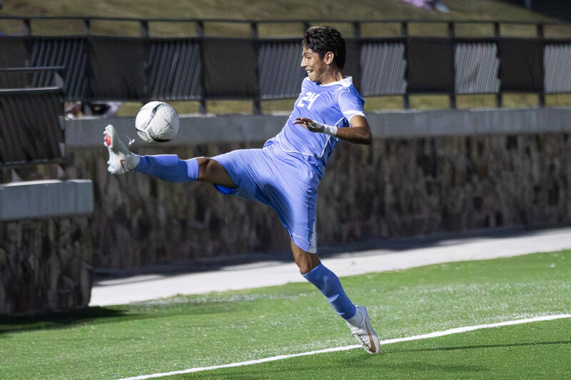 Soccer player in a light blue uniform jumps to control a ball mid-air by the sideline on a green field.