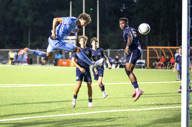 A soccer player in blue jumps to head the ball near the goal as defenders in dark uniforms watch during a night game.