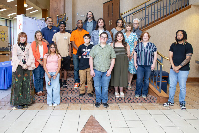 Fifteen diverse people smiling together on a staircase inside a building with tiled floors and decorative railing.