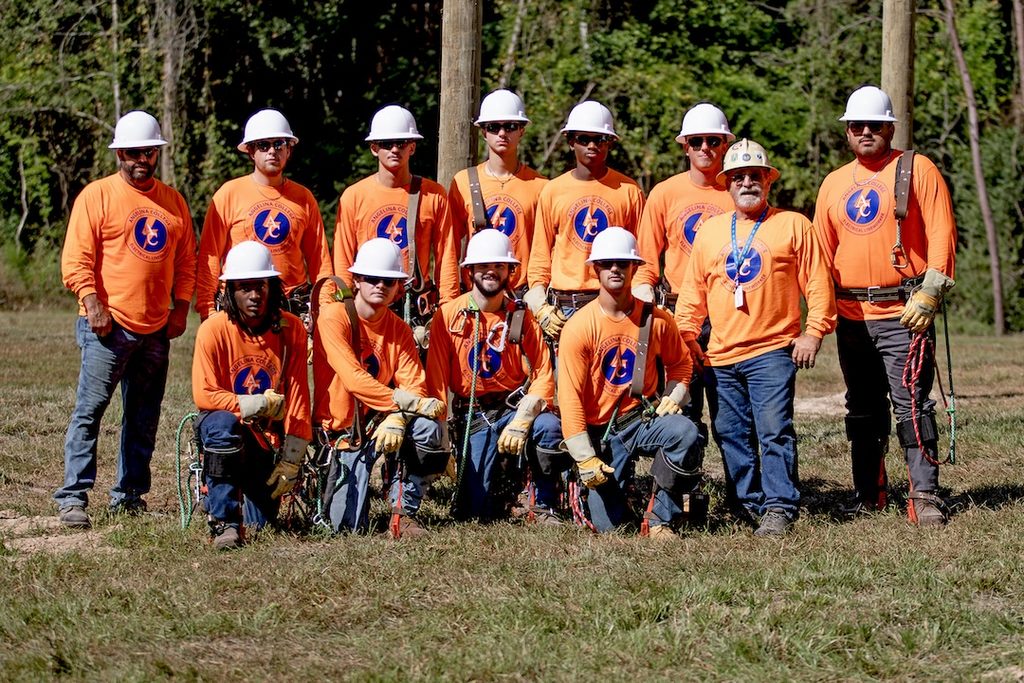 Twelve utility workers in orange shirts and hard hats pose on grass with trees behind; some kneel holding climbing gear and ropes.
