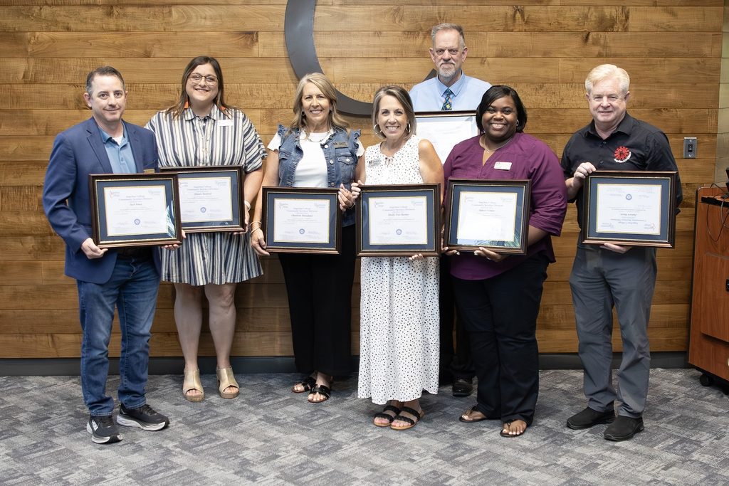 Seven people in business casual attire smile and hold framed certificates indoors, celebrating achievements by a wooden wall.