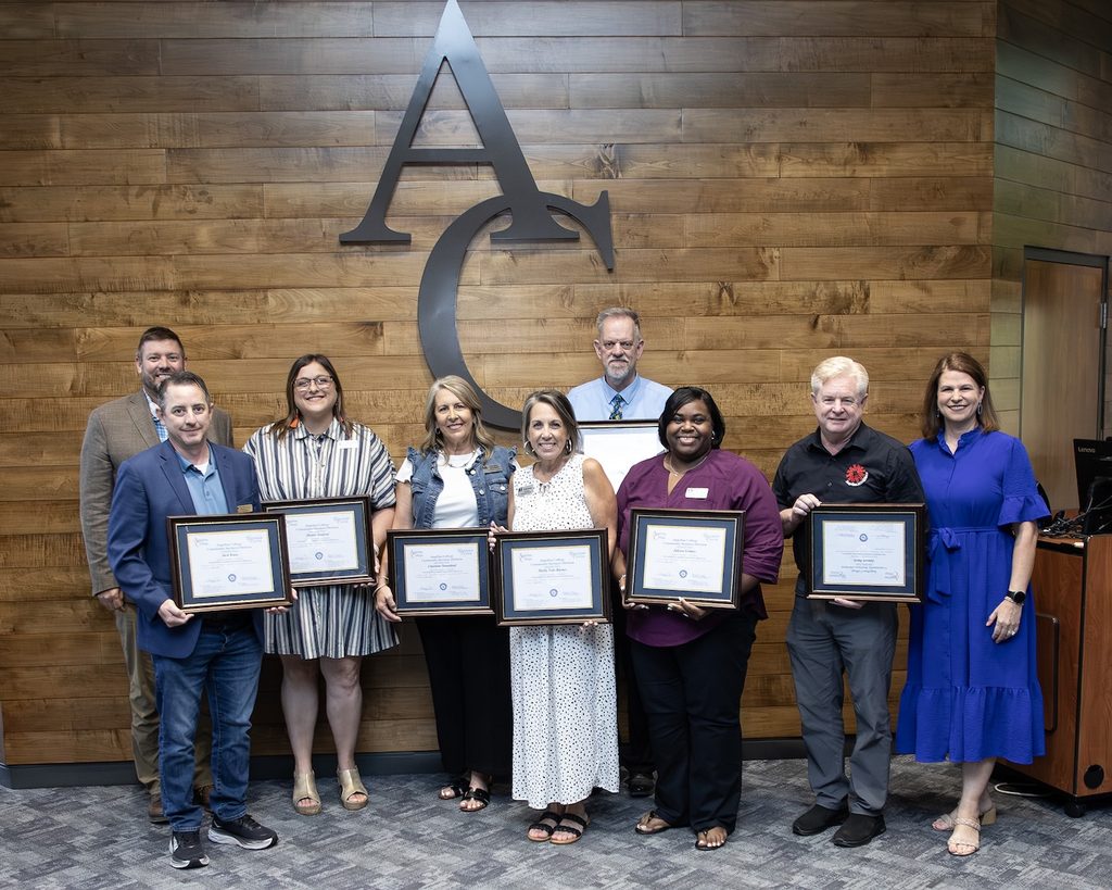 Eight people pose indoors, six holding framed certificates, smiling in business attire before a wooden wall with large AC letters.