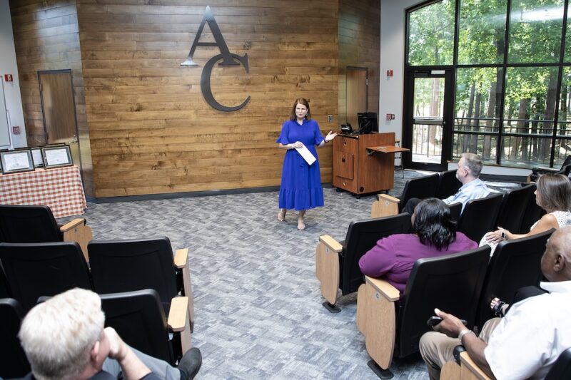 A woman in a blue dress speaks at a podium to a small audience in a modern room; “AC” is displayed on the wall behind her.