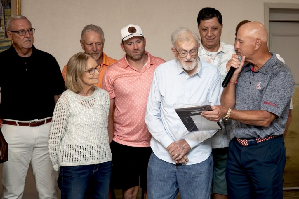 Several people stand indoors as a man with a microphone and plaque honors an elderly man in the center.