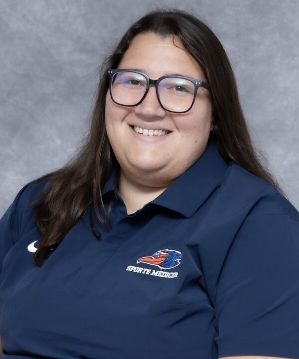 Smiling person with long dark hair and glasses, in a navy polo with bird logo and “SPORTS MEDICINE,” against gray background.