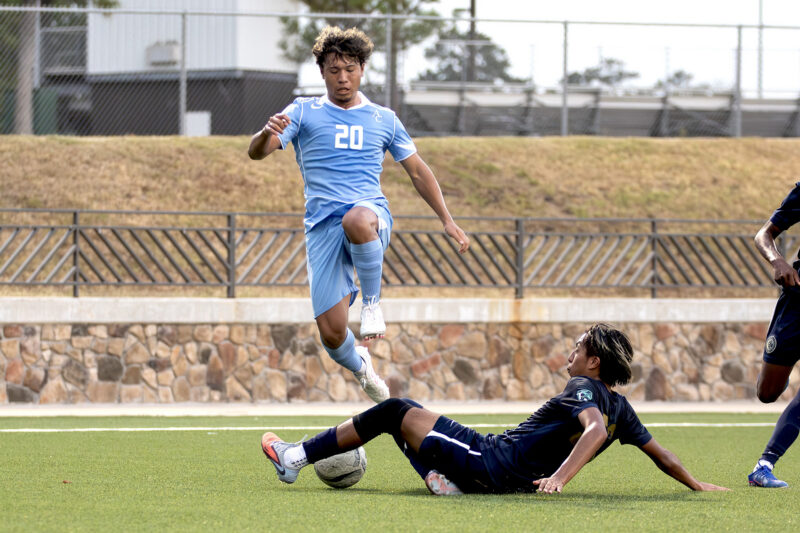 Soccer player in blue jersey jumps to dodge a sliding tackle from an opponent in navy on a grassy field.