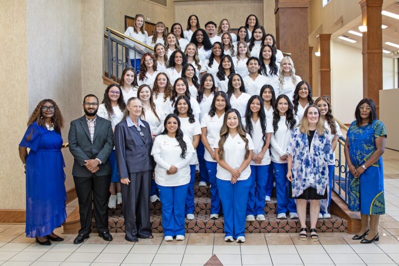 Nursing students in white and blue pose on indoor stairs with five faculty in formal wear; space is bright with wood accents.