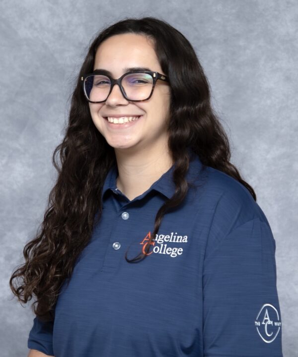 Person with long dark hair and glasses, smiling in a blue Angelina College polo, stands before a gray studio backdrop.